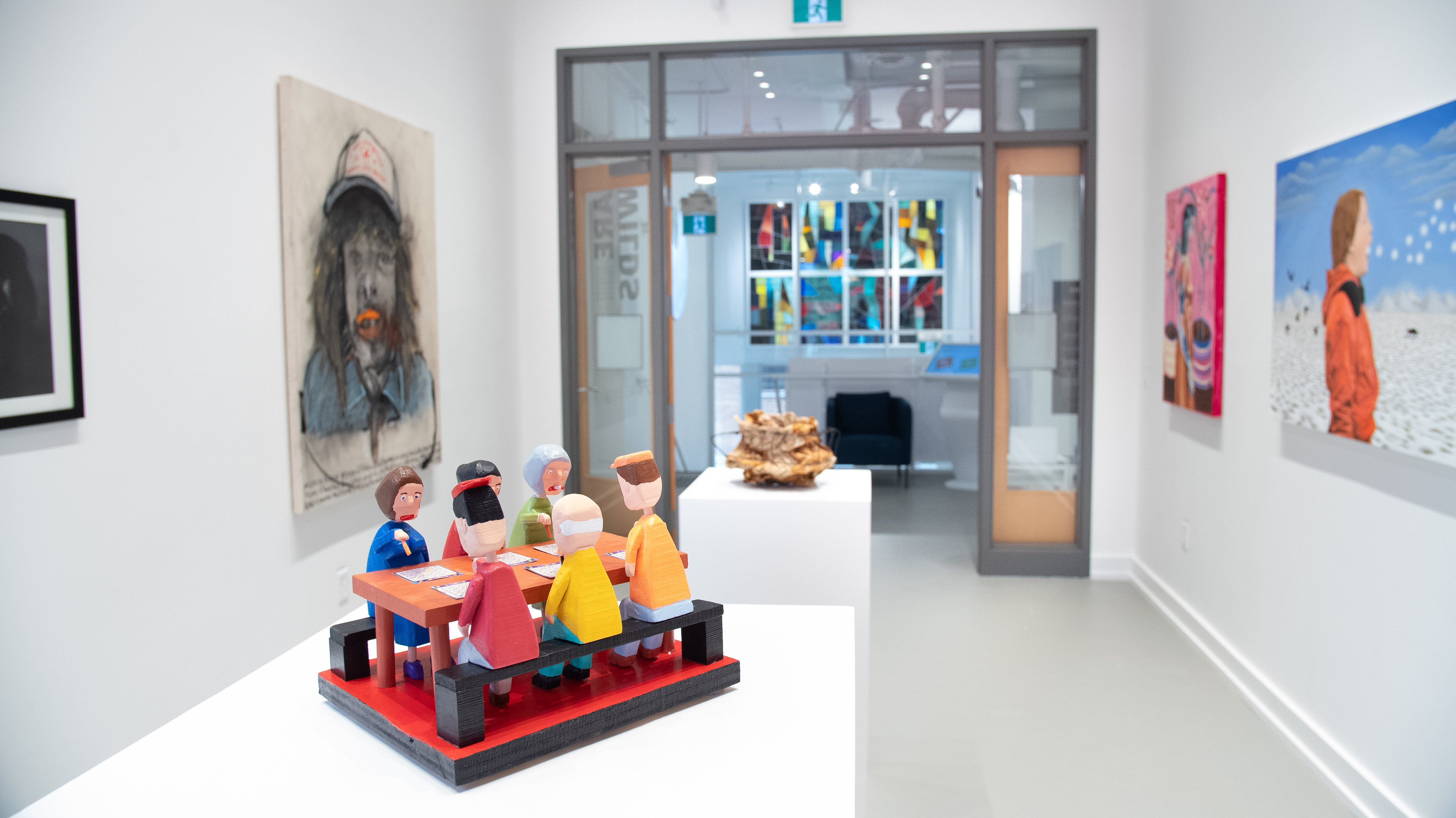 Are Wilds Exhibition. A white room with art on the walls and a focus on an art piece of six people at a table.